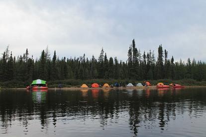 ue d’un campement de tentes colorées installées le long d'une rivière calme, avec une forêt dense de conifères en arrière-plan. Plusieurs canots et équipements sont disposés sur la rive.